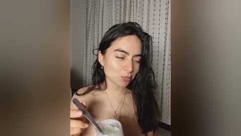 Media: A video of a young woman with long, wet, black hair, wearing a strapless top, eating ice cream with a spoon, indoors, against a patterned wall.