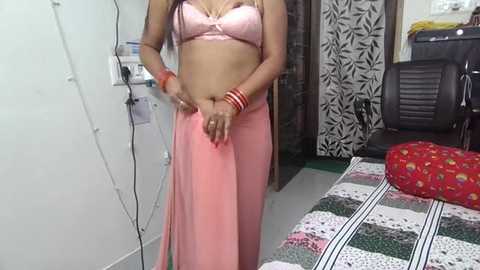 Video of a South Asian woman in a pink saree, standing in a modestly furnished bedroom with a floral-patterned bedspread and black and white patterned wall.