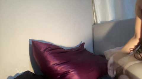 Video of a person's legs and torso in a dimly lit bedroom. The person is wearing a light-colored dress and sitting on a bed with a deep purple pillow and a beige blanket.