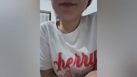 A video of a young woman with light skin, wearing a white T-shirt with the word \"Cherry\" in red, partially cropped. The background shows a white wall and a blurred white door.