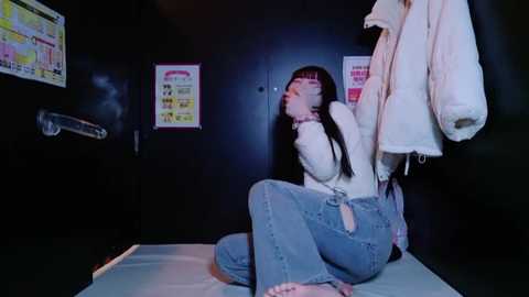 Video of a young woman in a dark room, kneeling with her hands tied behind her back, wearing a white jacket and blue jeans, a white coat hanging behind her.