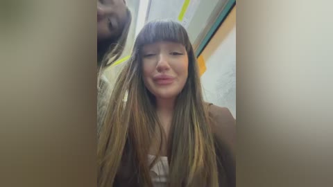 A video captures a young woman with long, straight, blonde hair and blunt bangs, wearing a sleeveless white top, smiling and sticking her tongue out. The background features a blurred, yellow-and-white-striped wall.