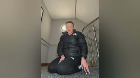 Media: Video of a fit, blonde woman with fair skin, kneeling on a carpeted floor in a dimly-lit room, wearing a black puffer jacket and leggings.