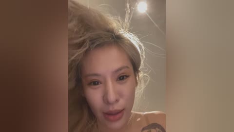 Video of a young Asian woman with light skin and messy, platinum blonde hair, wearing a light-colored top, smiling. Background shows a blurred, warm-toned room with a ceiling light.