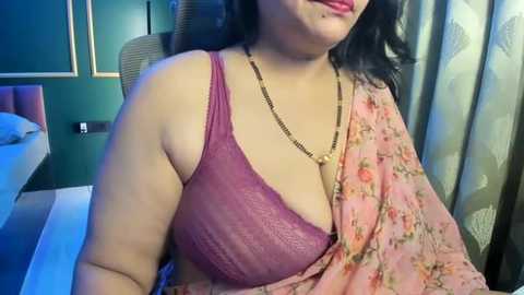 A video of a South Asian woman with medium skin tone, wearing a floral pink sari with a gold chain necklace. She has a voluptuous figure and dark, wavy hair.