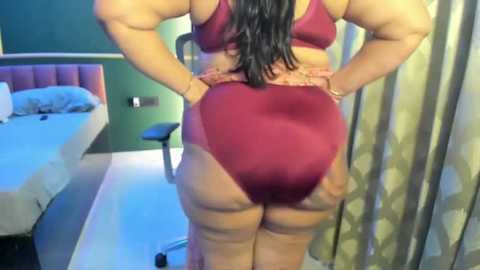 Video of a plus-sized woman in a maroon bra and thong, standing with hands on hips, facing away from camera. Background shows a bed, green wall, and patterned curtains.