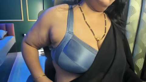 Video of a woman with medium skin tone, wearing a light blue bra and a black shawl, with long dark hair, in a bedroom with green walls, a bed, and a lamp.