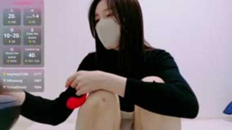 Video of an East Asian woman with long black hair and light skin wearing a black mask and long-sleeved shirt, seated with legs crossed, holding a red ball. Background features a digital screen displaying various game stats.