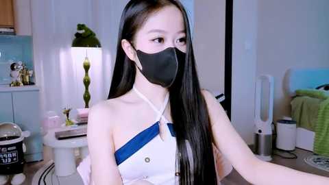 Media: Video of an East Asian woman with long black hair, wearing a black face mask, white dress, and seated in a modern, minimalist bedroom with a desk lamp, green plant, and white walls.