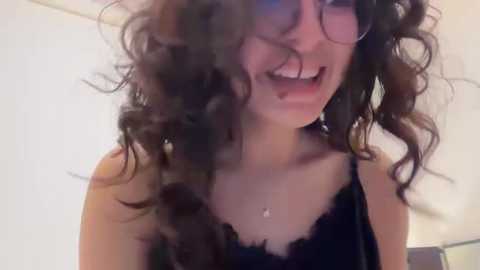 Media: Video of a young woman with curly, voluminous dark hair, laughing, wearing a black lace camisole, with a white background and visible wall.