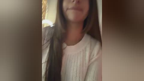 A video of a young woman with long, dark hair, wearing a light-colored, textured sweater, smiling slightly, partially out of focus, taken indoors with soft lighting.