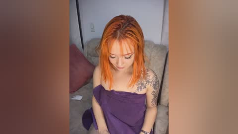 Video of a young woman with vibrant orange hair, wearing a purple towel, sitting on a beige couch, with visible tattoos on her left arm.