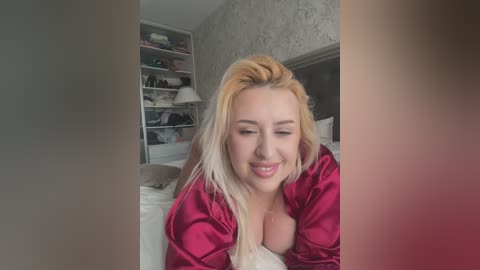 Video of a smiling, blonde-haired woman with fair skin, wearing a red satin robe, lying on a bed in a dimly lit bedroom with white wallpaper and shelves.