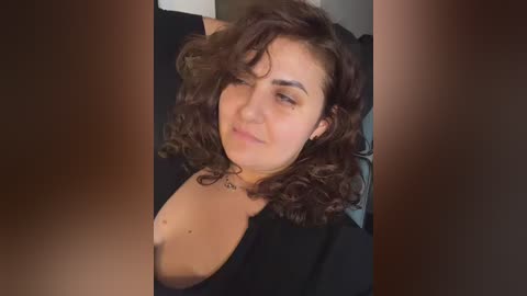 Video of a young, light-skinned woman with curly brown hair, wearing a black top, lying on a bed, smiling slightly. Background is blurred.