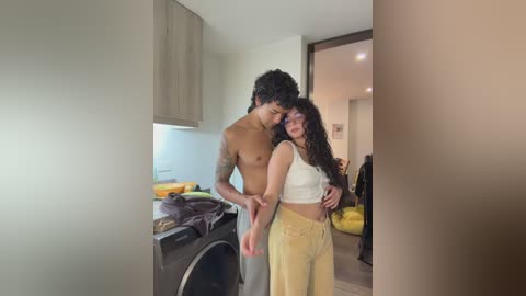 Media: Video of a shirtless, tattooed Black man and a curvy, light-skinned woman with curly hair in a modern, beige-toned kitchen. She wears a white crop top and yellow pants.