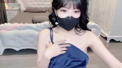 A video of an East Asian woman with shoulder-length black hair, wearing a black face mask, navy satin dress, and standing in a modern bedroom with white furniture.
