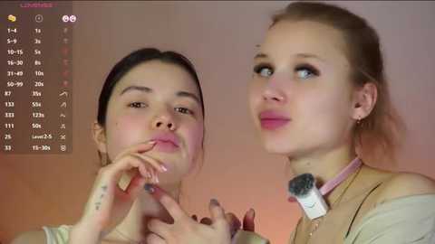 A video of two young women applying makeup, one with dark hair and the other with blonde hair, against a soft pink and orange background.