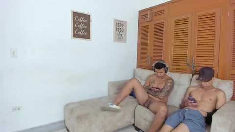 Media: Video of two shirtless men with tattoos lounging on a beige sectional couch, one in blue shorts, the other in a cap, looking at a smartphone. Wooden shuttered cabinets and wall art in a cozy living room.