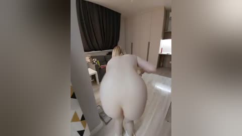 Video of a naked woman with fair skin and long blonde hair, bending over in a modern, minimally decorated bedroom with beige walls, dark curtains, and a geometric-patterned floor.