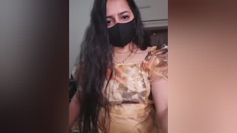 Video of a woman with long black hair, wearing a black mask, a yellow floral dress, and a gold necklace, standing indoors with a blurred background.