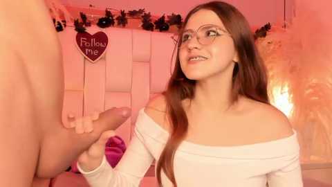 Media: Video of a young woman with glasses, long brown hair, and fair skin, smiling while holding a hard penis. Background includes a heart sign and warm, romantic lighting.