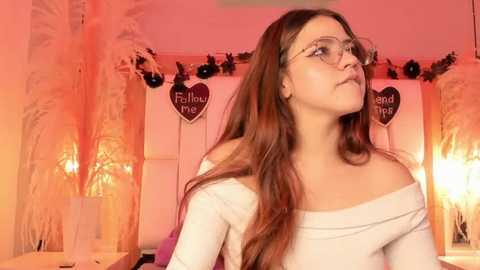 Media: A young woman with long brown hair and glasses stands in a warmly lit room, wearing a white off-shoulder top. Pink heart banners hang behind her.