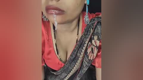 A close-up video of a woman in a red blouse and black sari, with a strand of drool on her lips, wearing blue earrings.