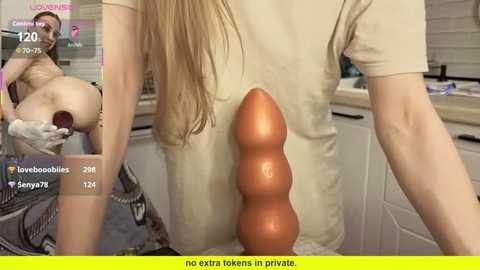 Video of a woman with long blonde hair, wearing a beige shirt, inserting a large, realistic brown dildo into her vagina in a kitchen.
