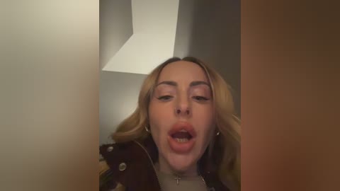 A candid video of a young woman with blonde hair, light skin, and a slight smile, wearing a black leather jacket, taken from a low angle, indoors with a white ceiling and brown wall.