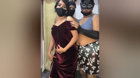 Video of three young women, two with dark skin and one with medium skin, wearing black face masks, burgundy velvet dresses, and white floral shorts, standing in a dimly lit, beige-walled room.