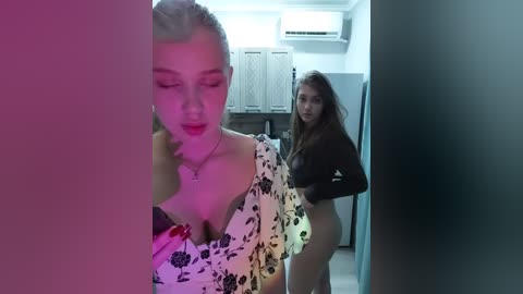 Video of two women: one blonde with a floral dress, another brunette in a black top, standing in a dimly lit kitchen with a pinkish-purple hue.