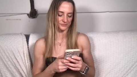 A video of a young woman with light skin and long blonde hair, wearing a strapless top, sitting on a white quilted couch, intently looking at her smartphone.