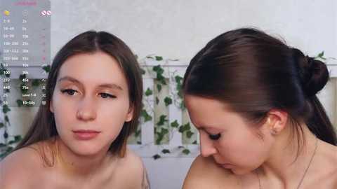 Video of two young women with pale skin, one with straight brown hair, the other with dark brown hair in a bun, both topless, sitting against a white wall with green ivy.