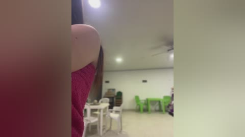 Video of a woman's upper arm in a pink sweater, viewed from behind, in a dimly lit, beige room with a ceiling fan, white chairs, and green plastic tables.