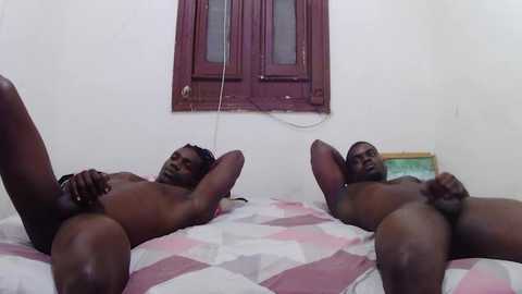 Video of two nude, muscular, black men with erect penises lying on a bed with a pink and white quilt. They have their legs spread apart, with one man's hand on his penis. The background features a maroon-framed window and a green book on a nightstand.