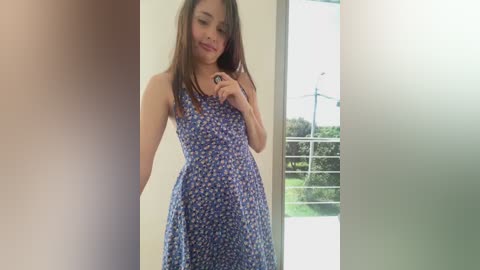 A video of a young woman with light skin and long brown hair, wearing a sleeveless, blue floral dress, standing indoors with a window behind her.