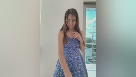 A video of a young woman with long brown hair, wearing a blue floral sundress, standing indoors near a large window with a scenic view of trees and a blue sky.