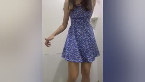 Video of a woman with light skin and long brown hair, wearing a sleeveless, blue and white floral-patterned dress, standing in a plain white-walled room with a small shelf and no visible furniture.