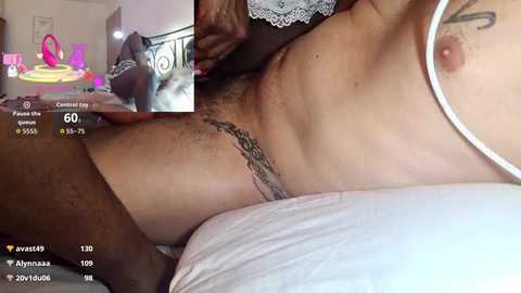 Video of a light-skinned woman with a tattoo on her hip, wearing white lace lingerie, lying on a bed with a man's arm visible.
