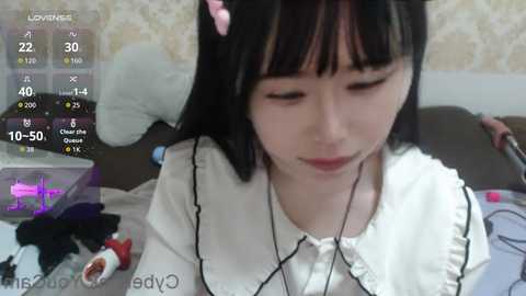 Video of an East Asian woman with long black hair, wearing a white frilled blouse, sitting on a bed, listening to headphones, in a cozy bedroom.