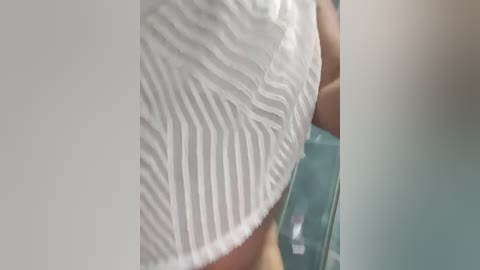 Media: Video of a woman's upper body wearing a white, ruffled, sleeveless top with a textured pattern. Her skin is light, and she appears to be indoors near a glass table.