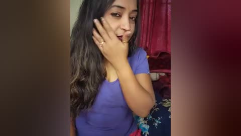 Media: A video of a young woman with long black hair, wearing a purple shirt, covering her mouth with her hand. She is indoors, near a red curtain and a colorful floral cloth.