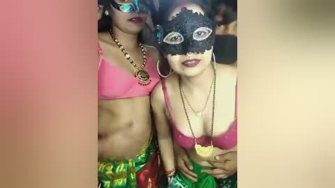 Media: Video of two women in vibrant, colorful costumes; one in a pink bikini, the other in a black mask, green skirt, and gold chain necklace.