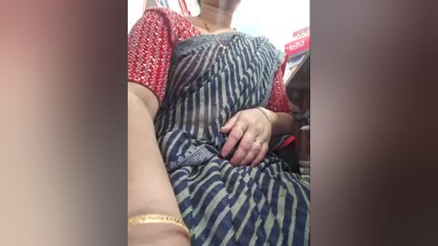 Video of a woman in a blue and white striped dress with red short sleeves, seated in a public space. She wears a gold bracelet on her left wrist. Background includes blurred objects.