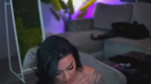 Video of a woman with dark hair, wearing a beige off-shoulder top, sitting on a couch. A black cat lies beside her, with a green plant and purple light in the background.