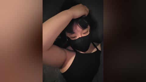 Media: A dimly-lit video shows a person with short black hair, wearing a black face mask and sleeveless black top, their arm raised to adjust their hair, partially obscured by a dark, shadowy background.