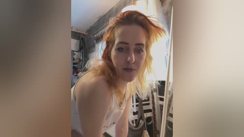 Media: Video of a pale-skinned, red-haired woman with messy hair, wearing a white tank top, leaning forward in a cluttered room.