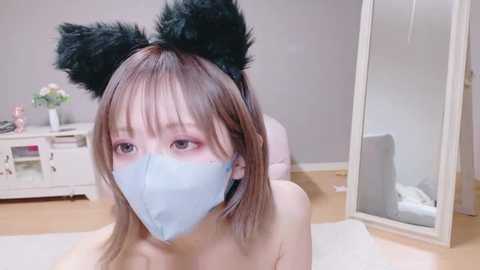 Media: Video of a young Asian woman with light skin, straight brown hair, wearing black cat ears and a light blue face mask, sitting on a white floor in a minimalist bedroom with white furniture and a mirror.