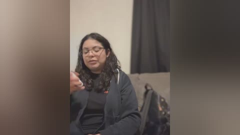 Video of a woman with long, curly black hair and glasses, wearing a black jacket and holding a phone, seated in a dimly lit room with beige walls and a black curtain.