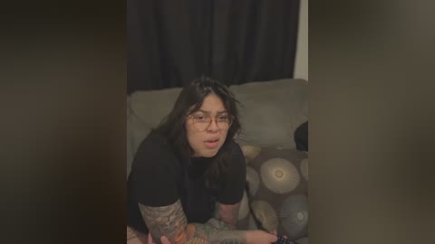 A video shows a woman with long, dark hair, wearing glasses and a black top, sitting on a couch with a patterned pillow. She has tattoos on her arm and appears to be looking off-camera with a neutral expression.
